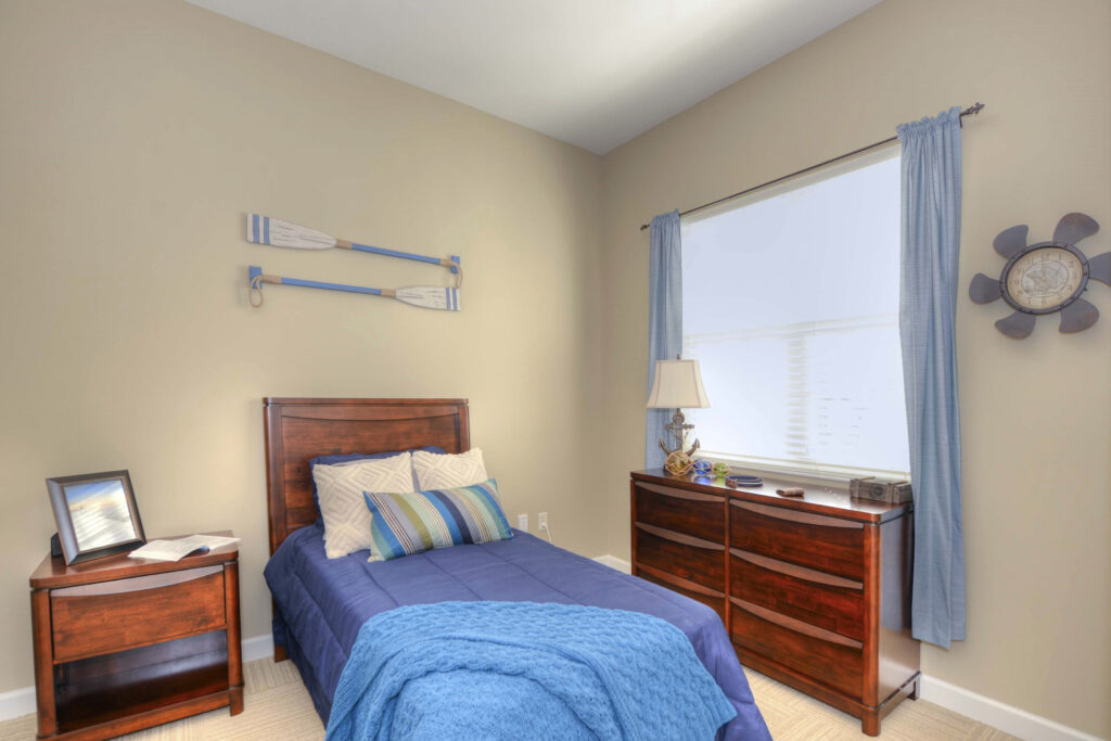 A bedroom with a small bed and blue sheets, throw pillows, a blue throw blanket, bedside table with a photo frame to the left of the bed, and a dresser with a lamp on it that sits underneath the window at Beach House, a senior living community.