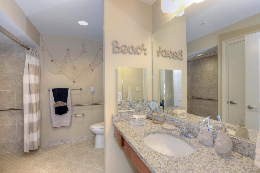 A bathroom with a marbled gray counter, mirror, sink, a walk-in shower, and a toile, Beach House senior living community.